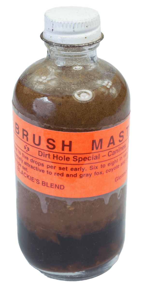 A small glass bottle labeled "Blackie's Brush Master Lure" is filled with brown liquid and sealed with a metal cap.